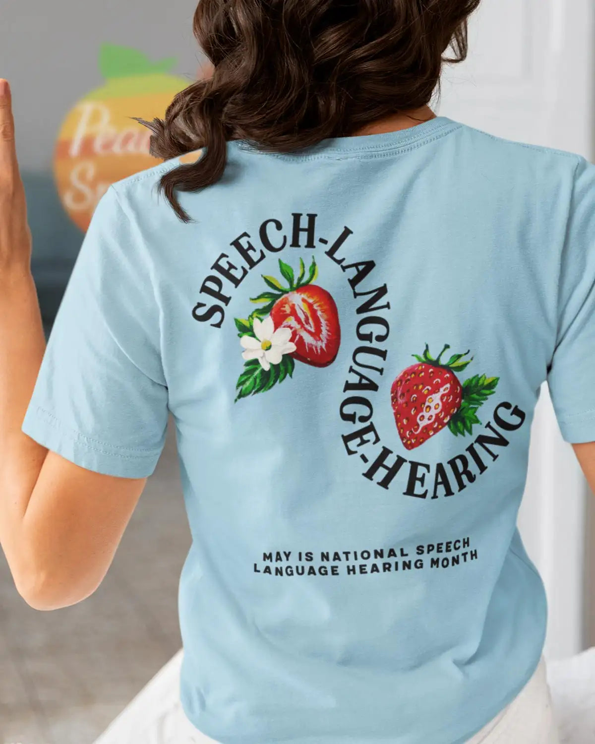 Light blue short-sleeved t-shirt with a circular graphic featuring strawberries and the text ’speech-language hearing’ arched above, along with smaller text below reading ’may is national speech language hearing month.’