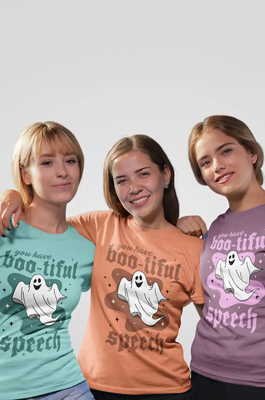 Three women wearing matching halloween-themed t-shirts featuring a smiling ghost graphic and the phrase ’you have boo-tiful speech’ in varying colors.